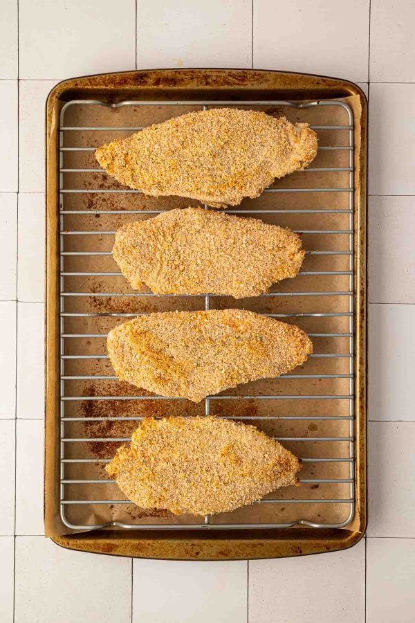 Four breaded chicken breasts arranged in a row on a wire rack over a baking sheet, ready to be cooked.