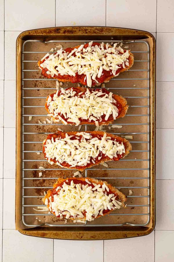 Four breaded chicken breasts topped with tomato sauce and shredded cheese on a wire rack over a baking sheet, ready to be baked.