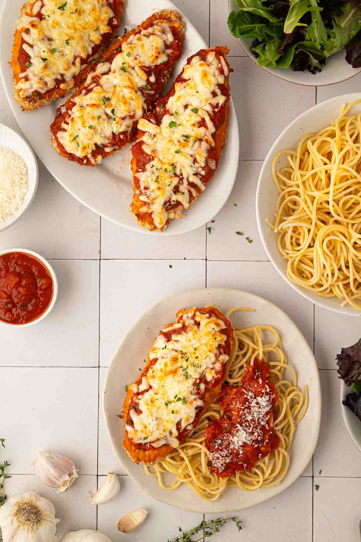 Plate of chicken parmesan with melted cheese served over spaghetti, surrounded by extra chicken, a bowl of spaghetti, tomato sauce, grated cheese, salad, and garlic on a white tile surface.