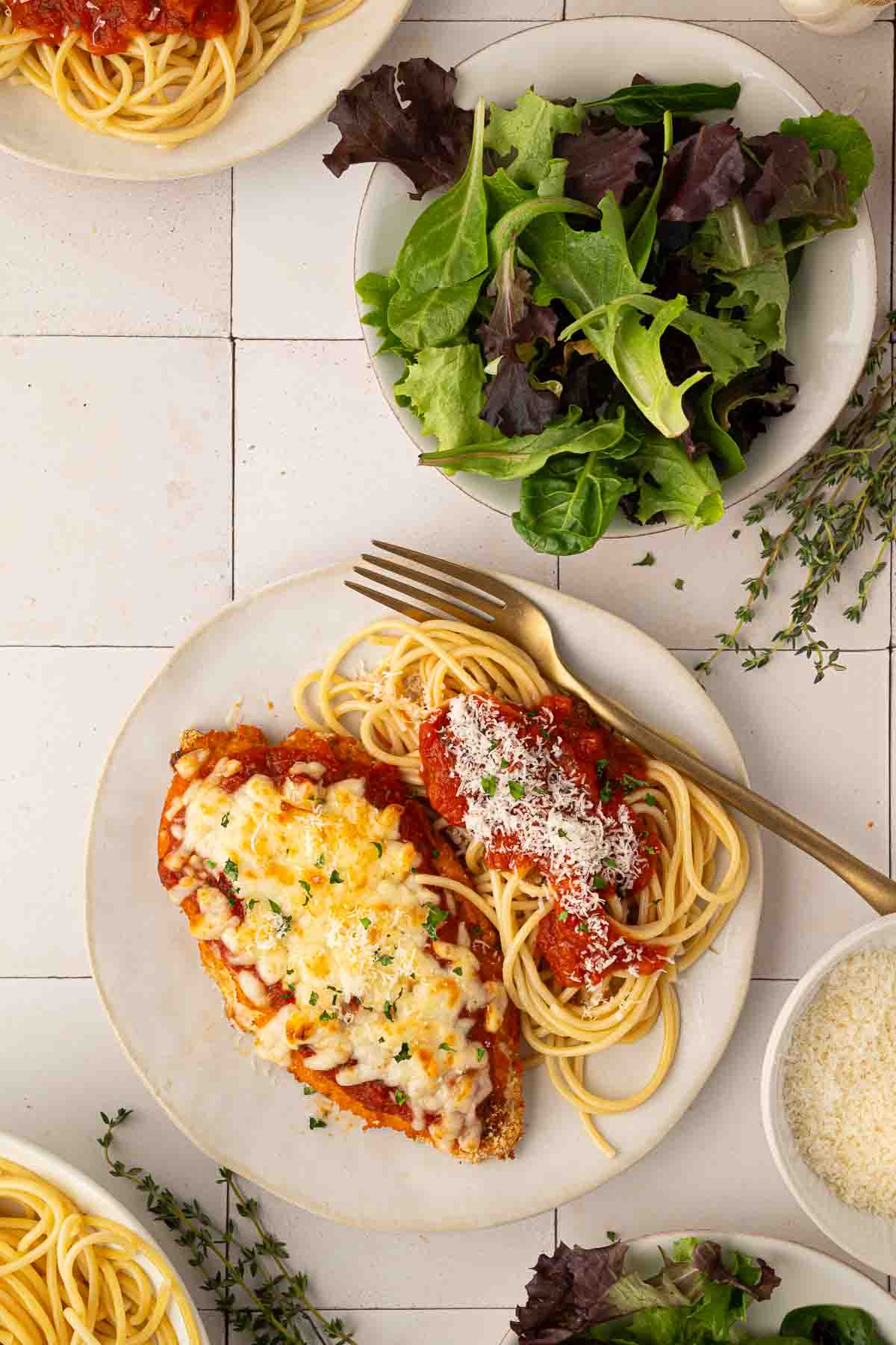 A plate with breaded chicken topped with melted cheese and tomato sauce, served alongside spaghetti with tomato sauce and grated cheese; a bowl of mixed greens is nearby.