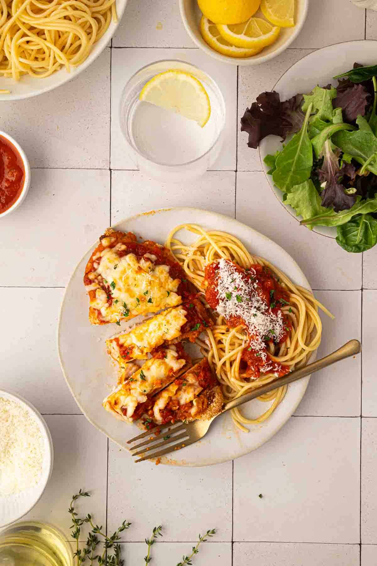 Plate with chicken parmesan topped with melted cheese, spaghetti with tomato sauce and grated cheese, and a fork. Surrounding dishes include salad, lemon slices, and pasta.