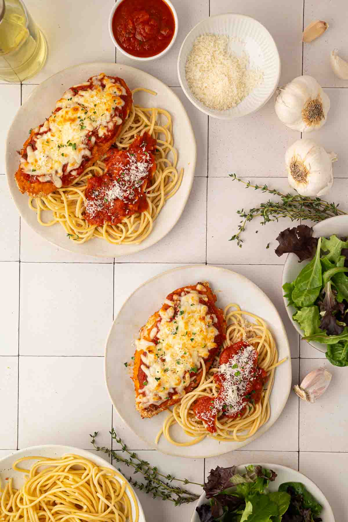 Two plates of spaghetti topped with chicken parmesan, garnished with herbs and cheese, are on a tiled surface alongside bowls of salad, grated cheese, tomato sauce, garlic, and fresh herbs.
