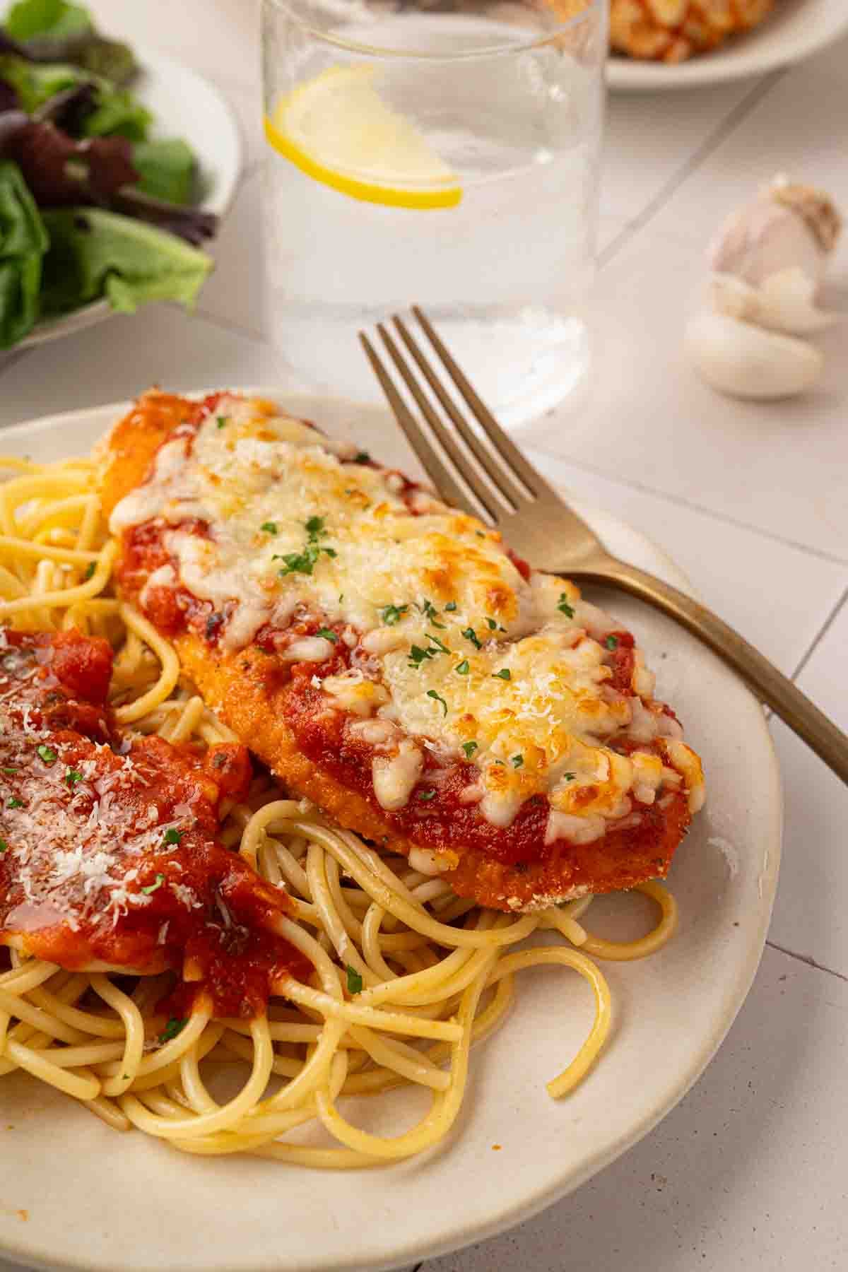 A plate of spaghetti with tomato sauce and breaded chicken parmesan topped with melted cheese, served with a fork. A glass of water with lemon and salad are in the background.