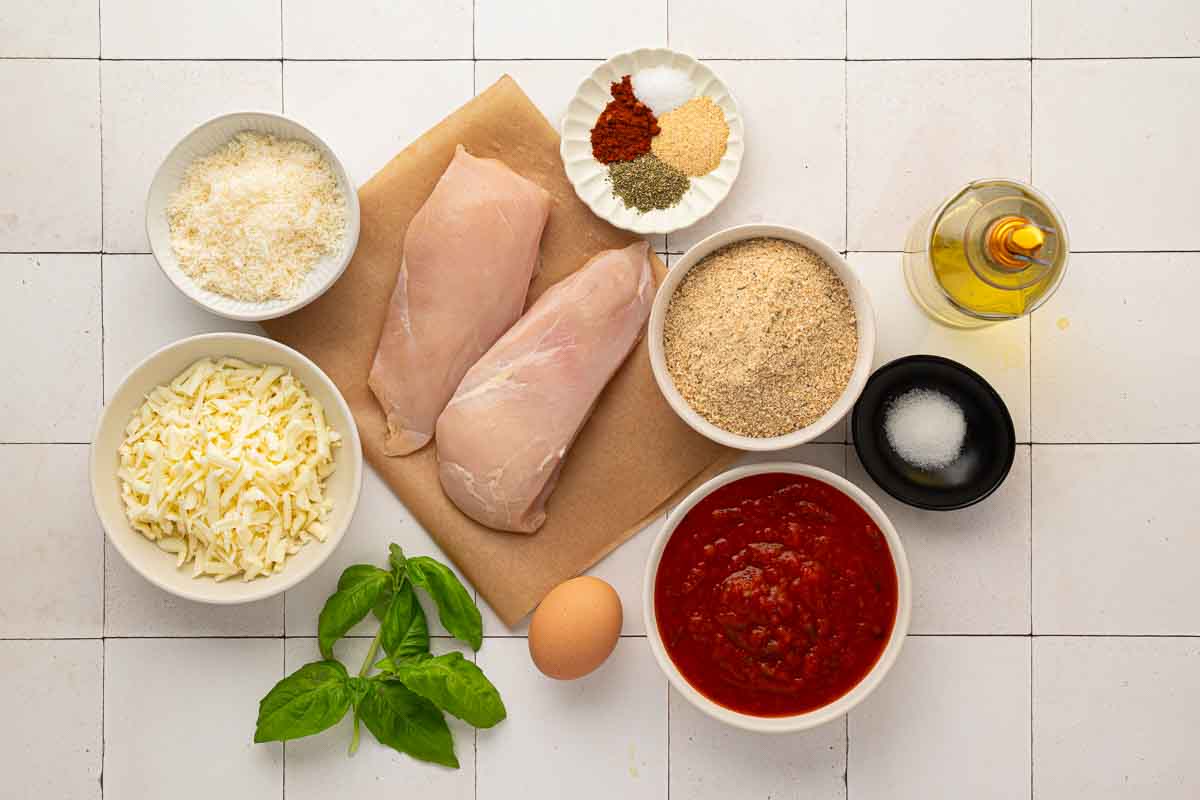 Ingredients for chicken parmesan arranged on a white tiled surface, including raw chicken breasts, cheese, breadcrumbs, marinara sauce, an egg, oil, spices, salt, and fresh basil.