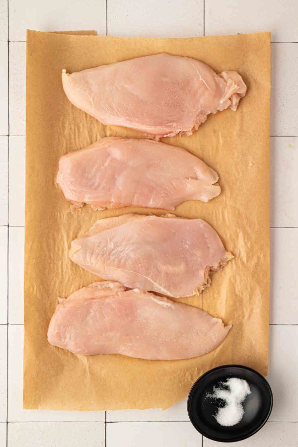 Four raw chicken breasts are arranged on parchment paper, with a small black bowl of salt placed nearby on a white tiled surface.