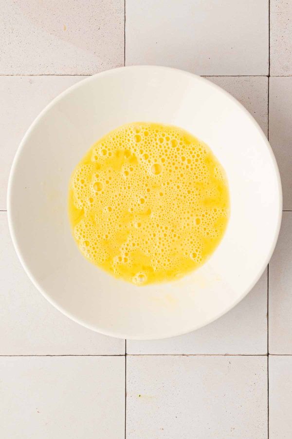 A white bowl containing whisked eggs with bubbles on the surface, placed on a tiled countertop.