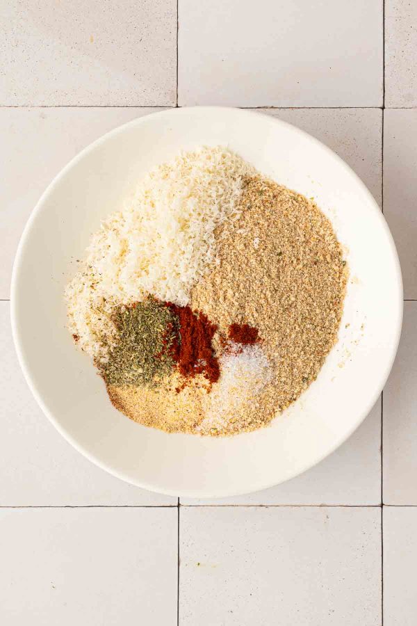 A white bowl containing shredded cheese, breadcrumbs, and assorted spices sits on a white tiled surface.