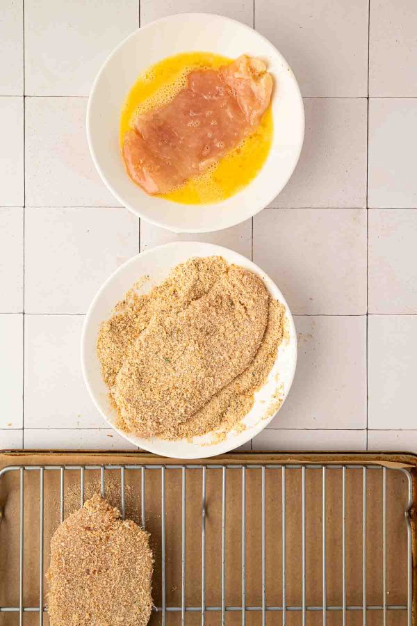 A raw chicken cutlet in beaten egg, another coated in breadcrumbs, and a breaded cutlet on a wire rack over a baking sheet.