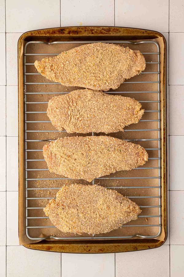 Four breaded chicken breasts are arranged in a single layer on a wire rack set over a parchment-lined baking sheet.
