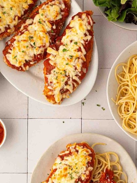 Sheet pan chicken parmesan topped with melted cheese and tomato sauce on plates, served with spaghetti, marinara sauce, grated cheese, and a side of mixed greens.