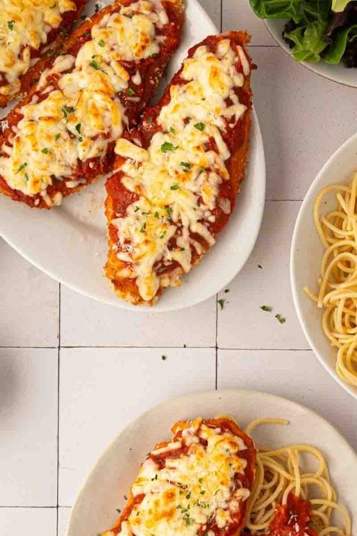 Sheet pan chicken parmesan topped with melted cheese and tomato sauce on plates, served with spaghetti, marinara sauce, grated cheese, and a side of mixed greens.