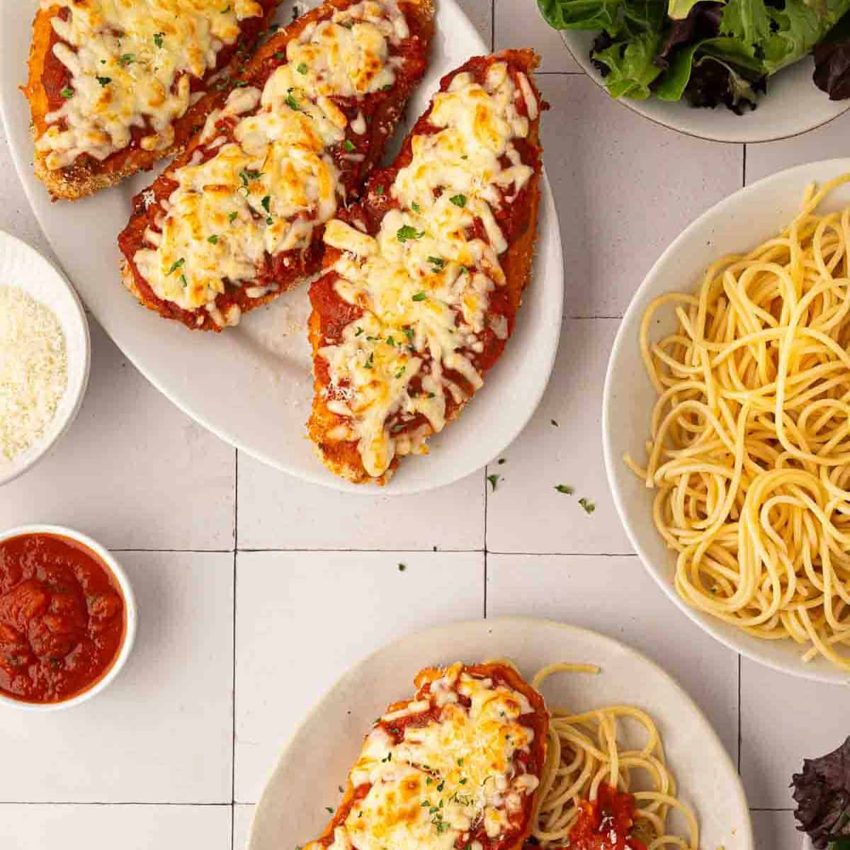 Sheet pan chicken parmesan topped with melted cheese and tomato sauce on plates, served with spaghetti, marinara sauce, grated cheese, and a side of mixed greens.