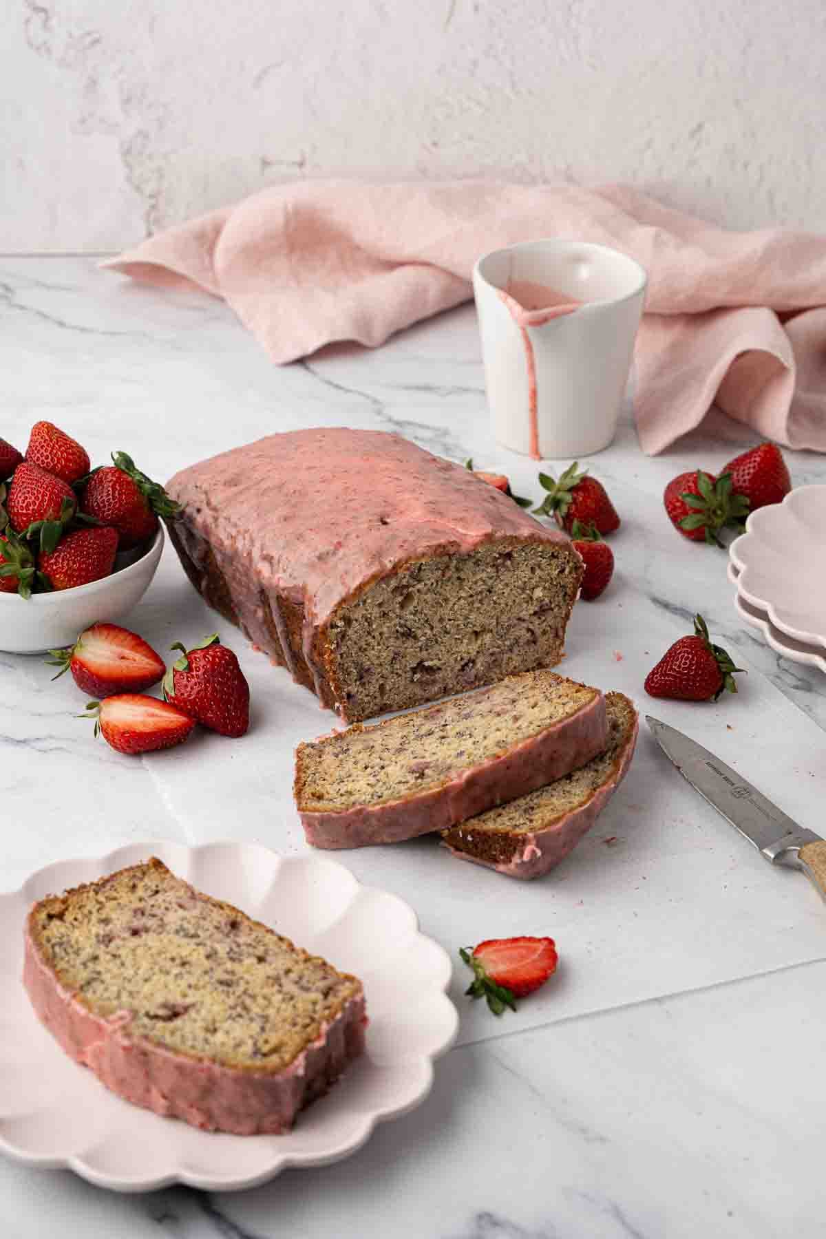 A loaf of strawberry banana bread with pink glaze, partially sliced on a plate, surrounded by fresh strawberries, a knife, and a cup with pink liquid on a marble surface.