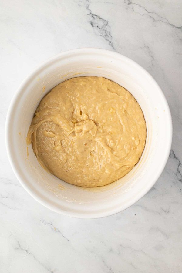 A white mixing bowl containing thick, light brown batter sits on a marble countertop.
