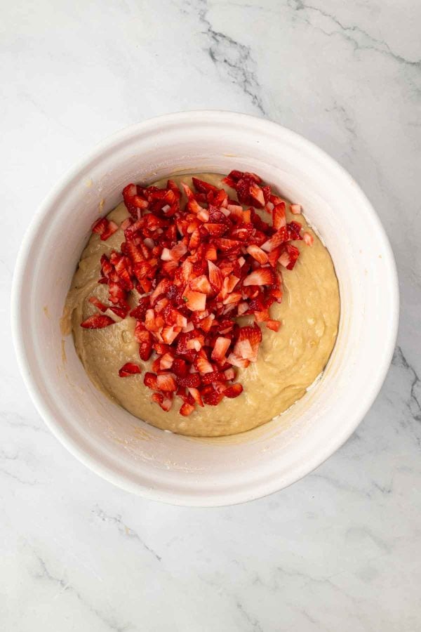 White bowl with cake batter topped with chopped fresh strawberries, sitting on a marble countertop.