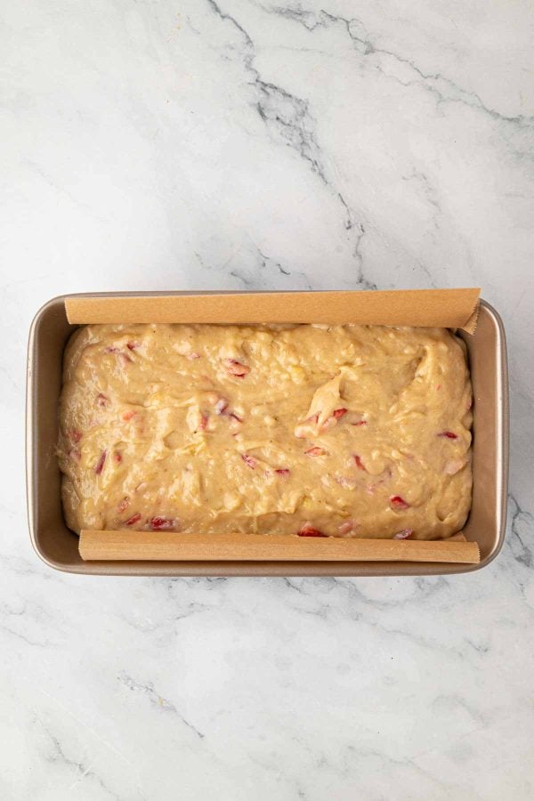 A loaf pan lined with parchment paper is filled with unbaked batter containing visible pieces of fruit, set on a white marble surface.