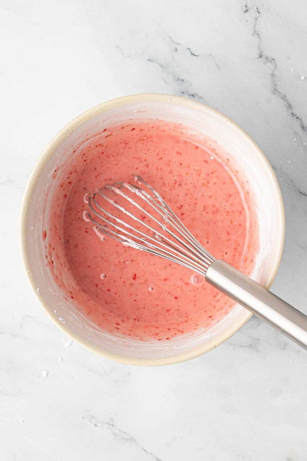 A mixing bowl filled with pink batter and a metal whisk on a marble countertop.