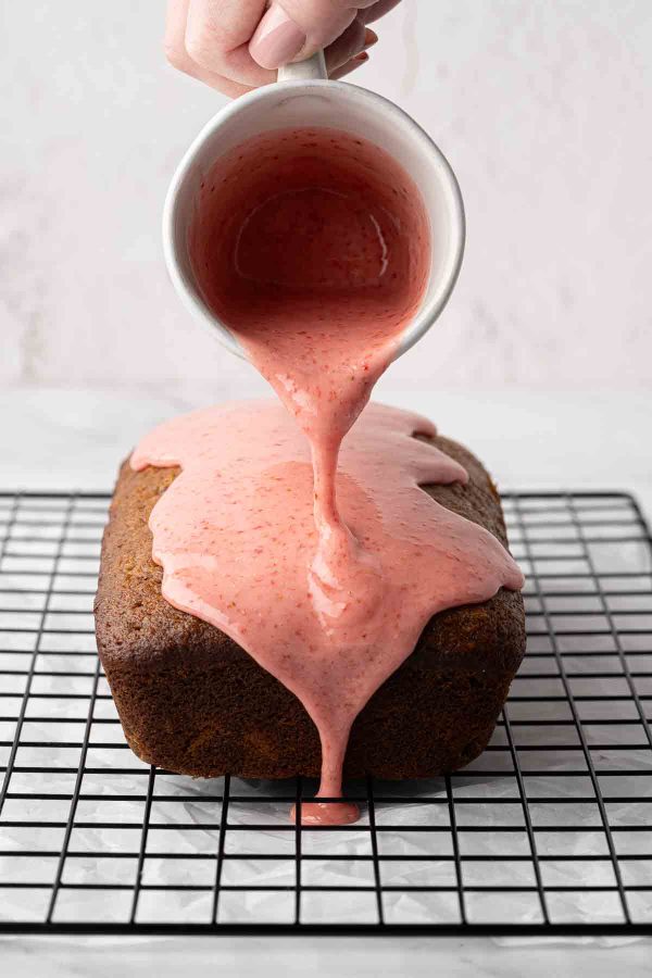A hand pours pink strawberry glaze from a small pitcher onto a loaf strawberry banana bread placed on a black wire cooling rack.