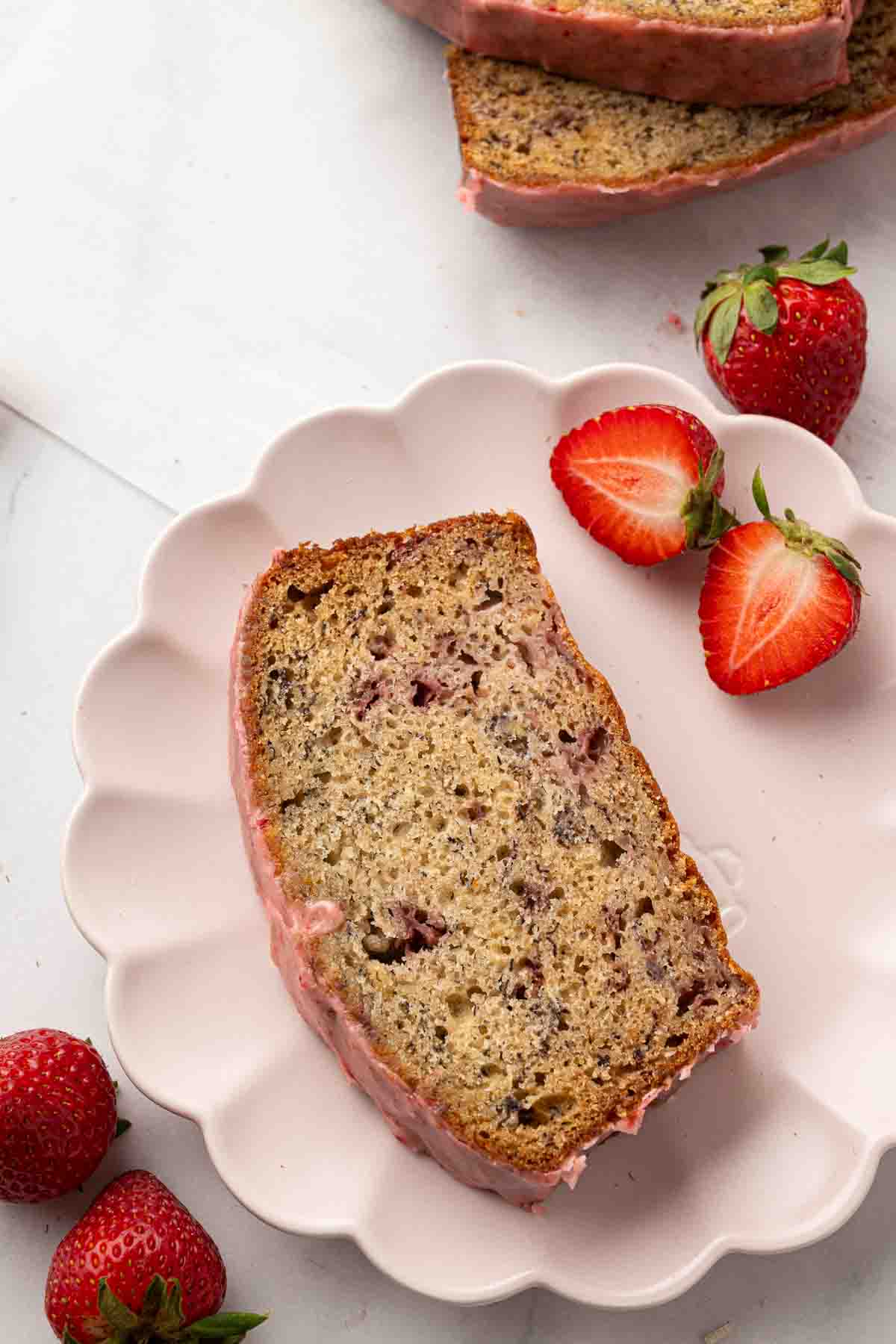 A slice of strawberry banana bread with pink glaze on a white plate, surrounded by fresh strawberries and strawberry halves.