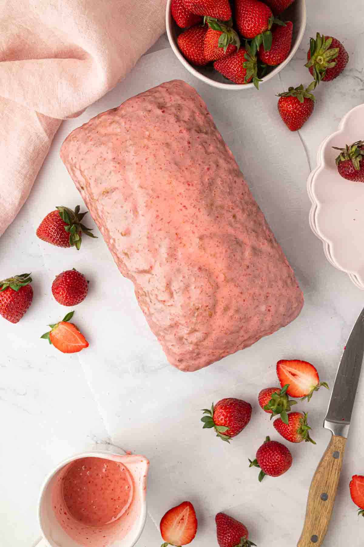 A loaf cake covered in pink strawberry glaze is surrounded by fresh strawberries, a knife, a bowl of strawberries, and a small pitcher of pink glaze on a white surface.