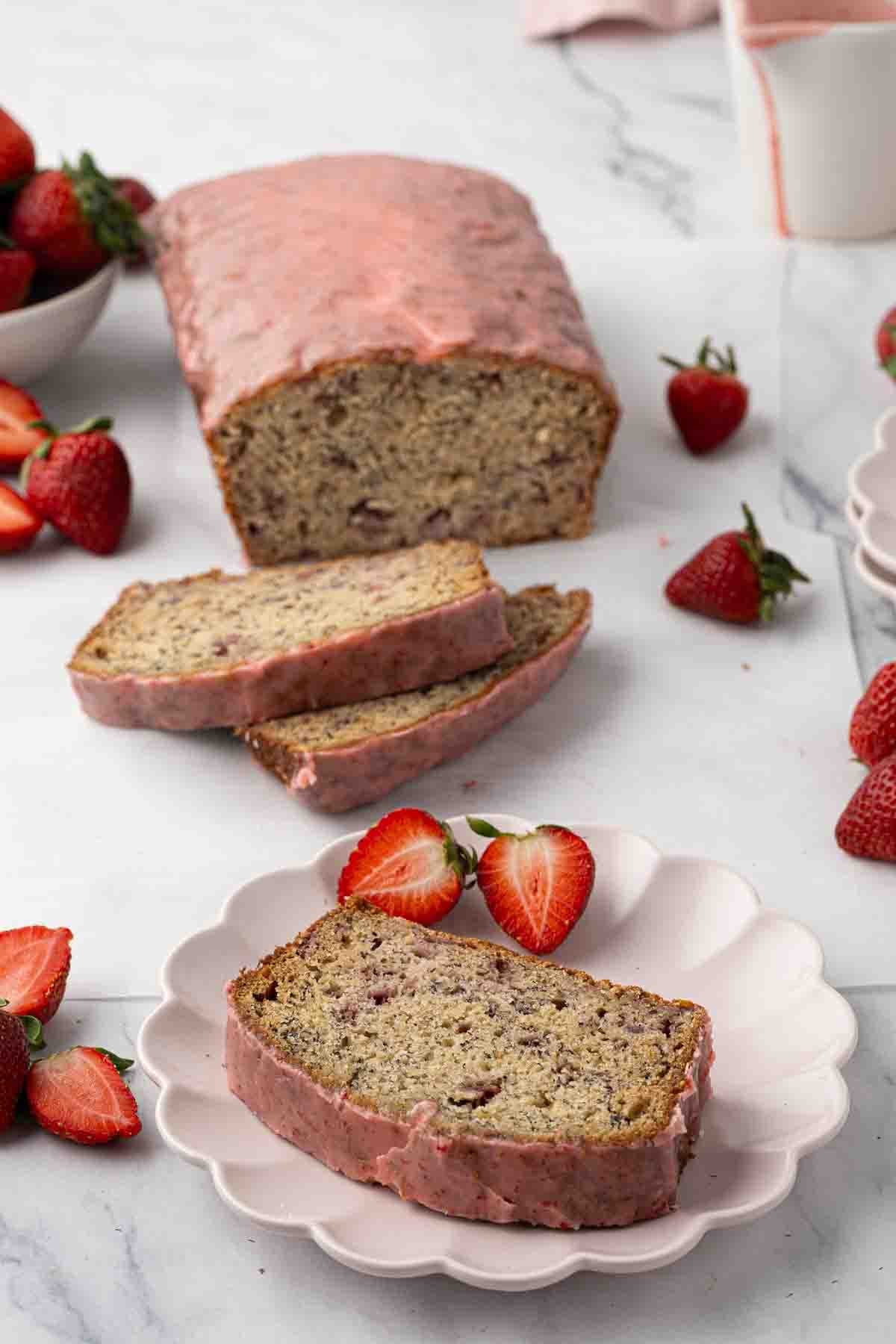 A loaf of banana bread with pink strawberry glaze, two slices cut, one placed on a plate with halved strawberries. Whole strawberries are scattered around on a marble surface.