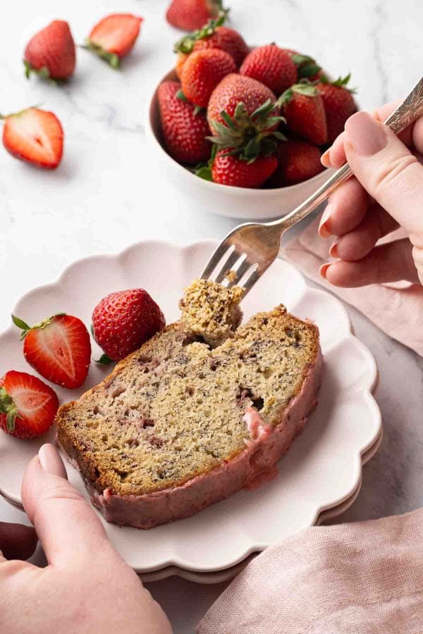 A hand holds a fork over a slice of strawberry banana bread with strawberries on a plate, with a bowl of whole strawberries in the background.