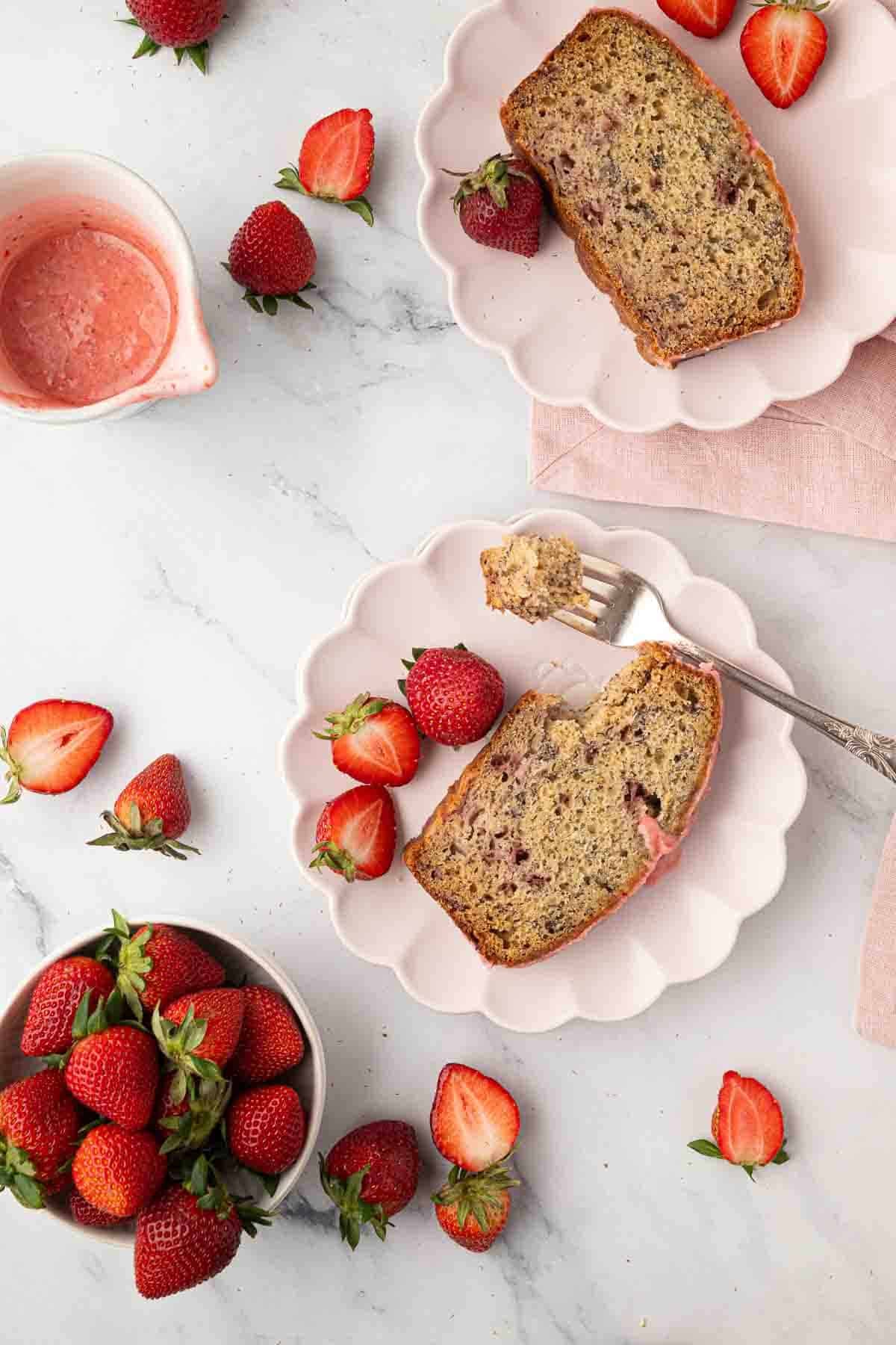 Two plates each with a slice of strawberry banana bread and strawberries on a white surface, surrounded by more strawberries and a small pitcher of pink sauce.