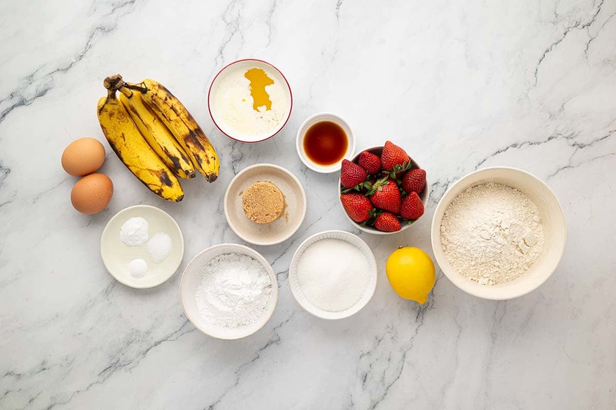 A variety of baking ingredients are arranged on a marble surface, including eggs, bananas, strawberries, flour, sugar, brown sugar, lemon, vanilla, oil, and powders.