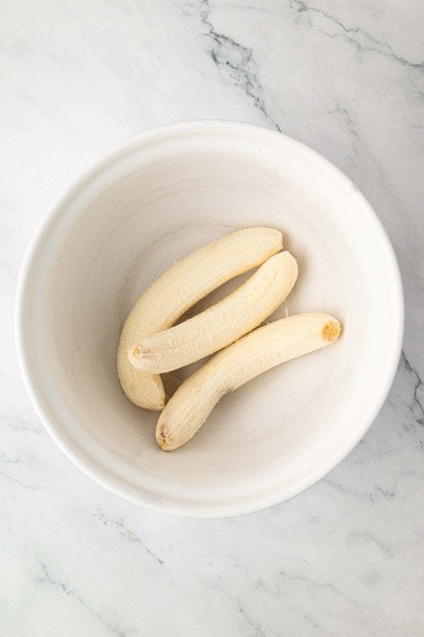 Three peeled bananas placed in a large white mixing bowl on a light marble countertop.