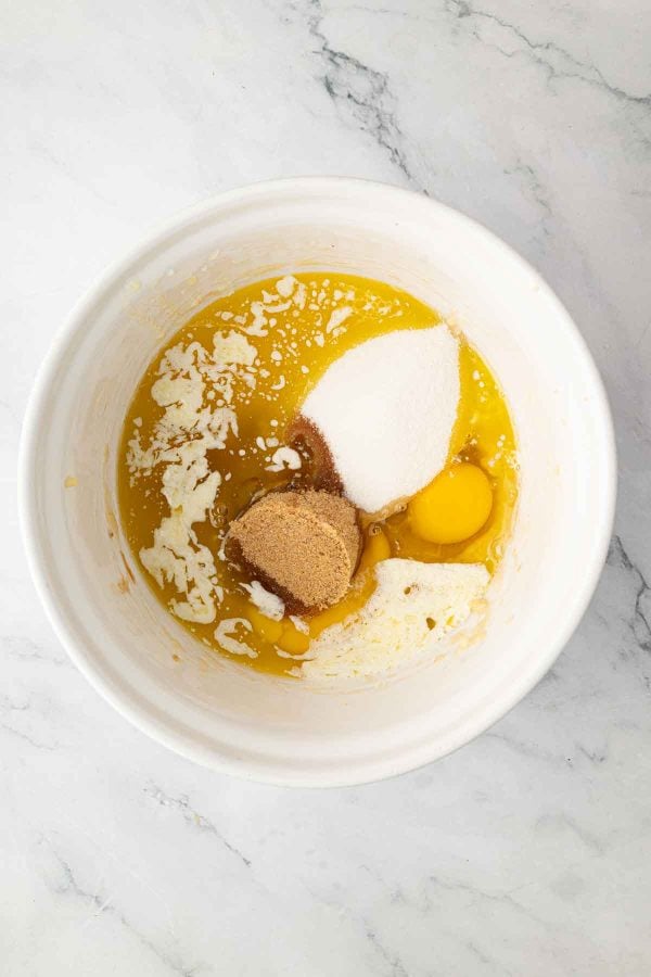 A white mixing bowl containing melted butter, granulated sugar, brown sugar, an egg, and milk on a marble countertop.