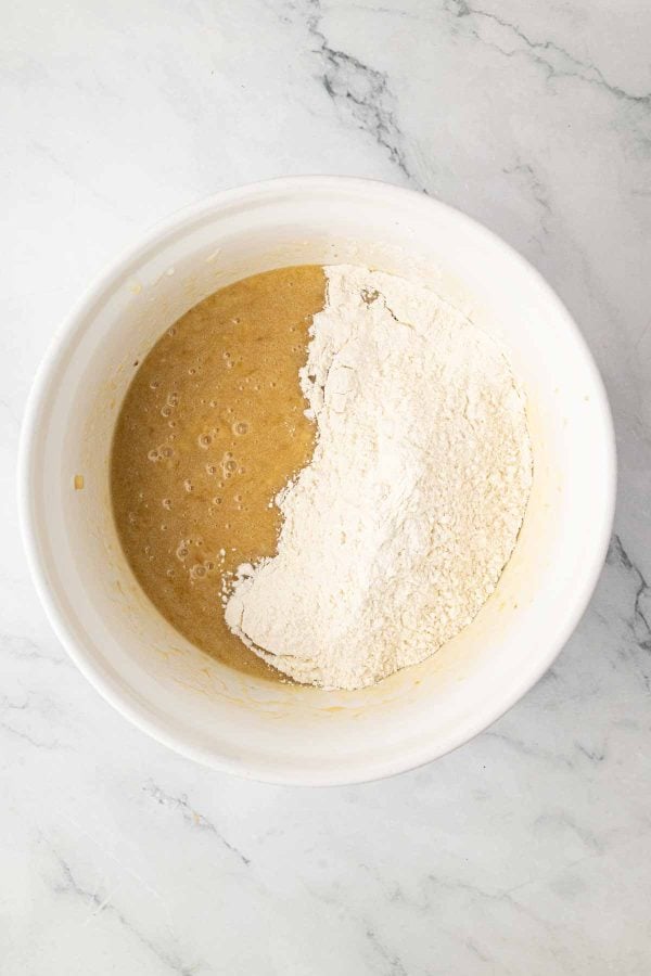 A white mixing bowl containing a brown liquid mixture and a mound of white flour on a marble surface.