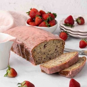A loaf of strawberry banana bread with pink strawberry glaze is sliced on a white surface, surrounded by fresh strawberries, a bowl of strawberries, a stack of plates, and a knife.