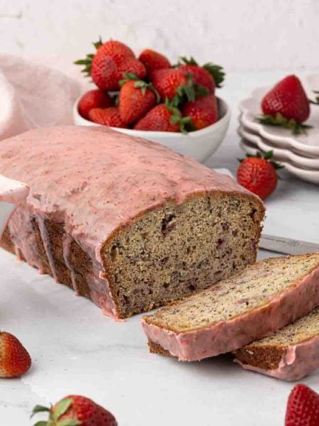 A loaf of strawberry banana bread with pink strawberry glaze is sliced on a white surface, surrounded by fresh strawberries, a bowl of strawberries, a stack of plates, and a knife.
