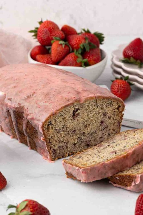 A loaf of strawberry banana bread with pink strawberry glaze is sliced on a white surface, surrounded by fresh strawberries, a bowl of strawberries, a stack of plates, and a knife.