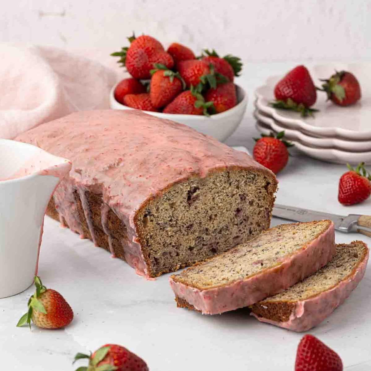 A loaf of strawberry banana bread with pink strawberry glaze is sliced on a white surface, surrounded by fresh strawberries, a bowl of strawberries, a stack of plates, and a knife.
