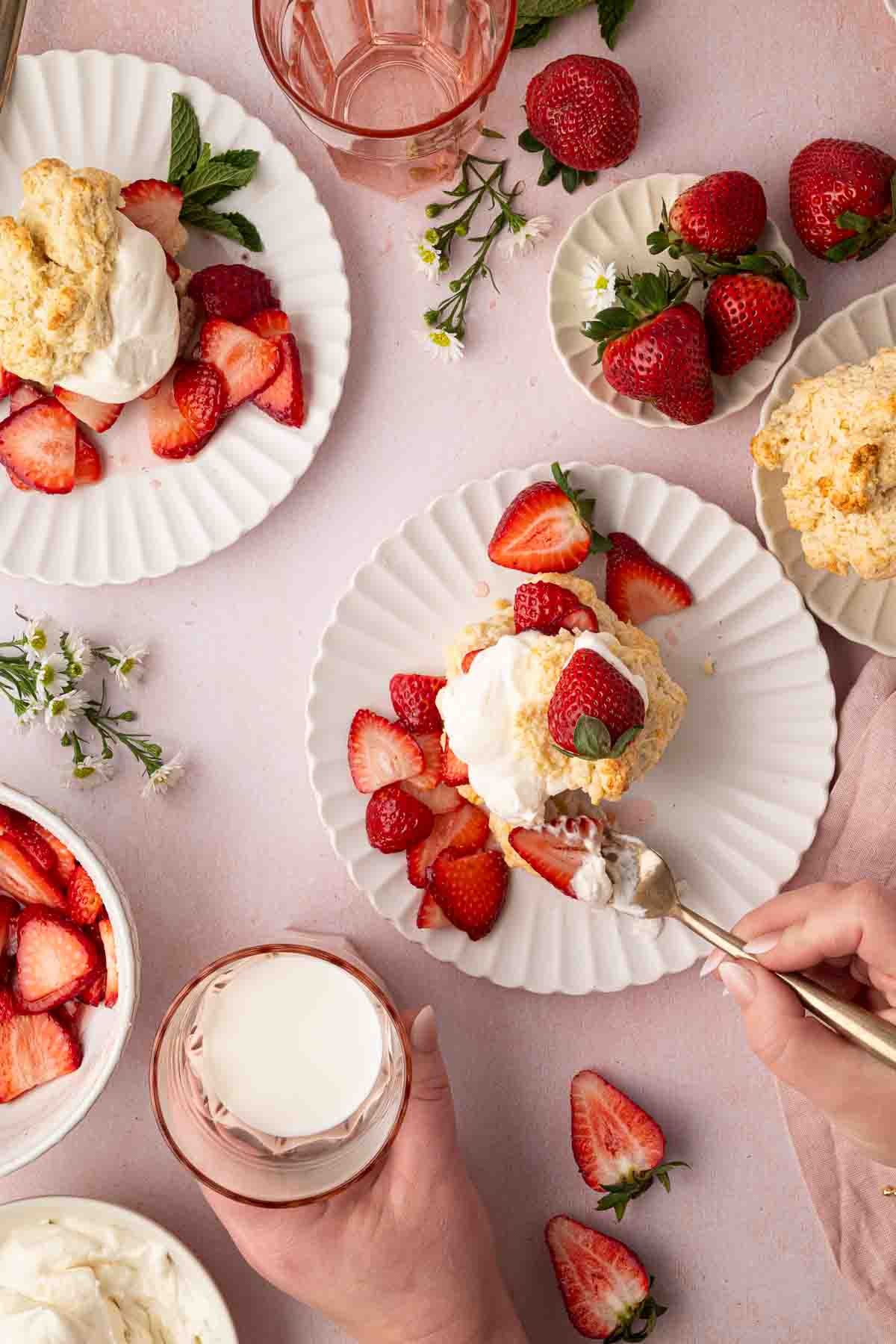 A table set with plates of strawberry shortcake topped with whipped cream, fresh strawberries, a glass of milk, and scattered strawberry halves on a light pink surface.