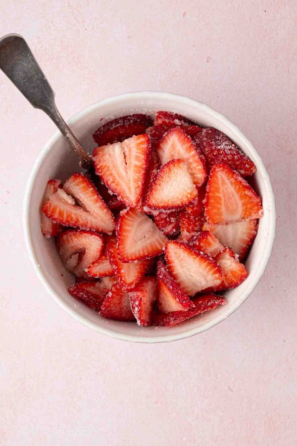A bowl of sliced strawberries sprinkled with sugar, with a spoon resting inside, on a light pink surface.