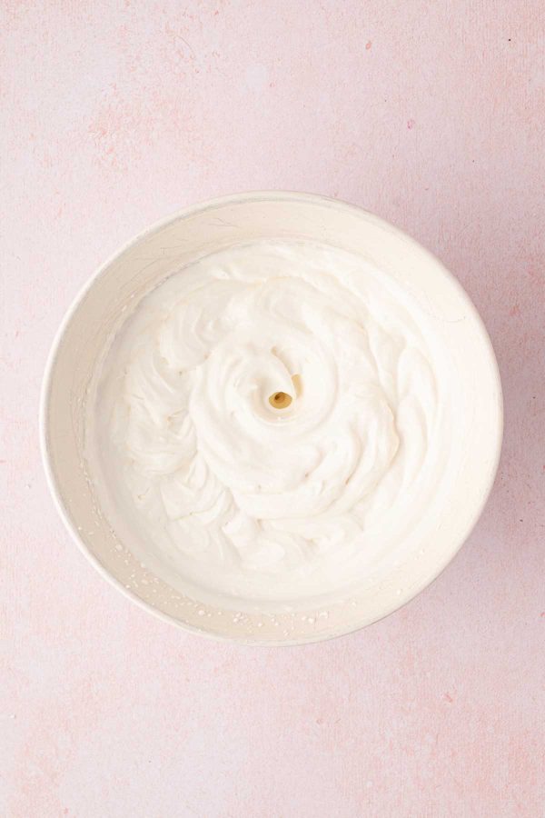 A bowl of whipped cream with a whisk hole in the center, set on a light pink surface.