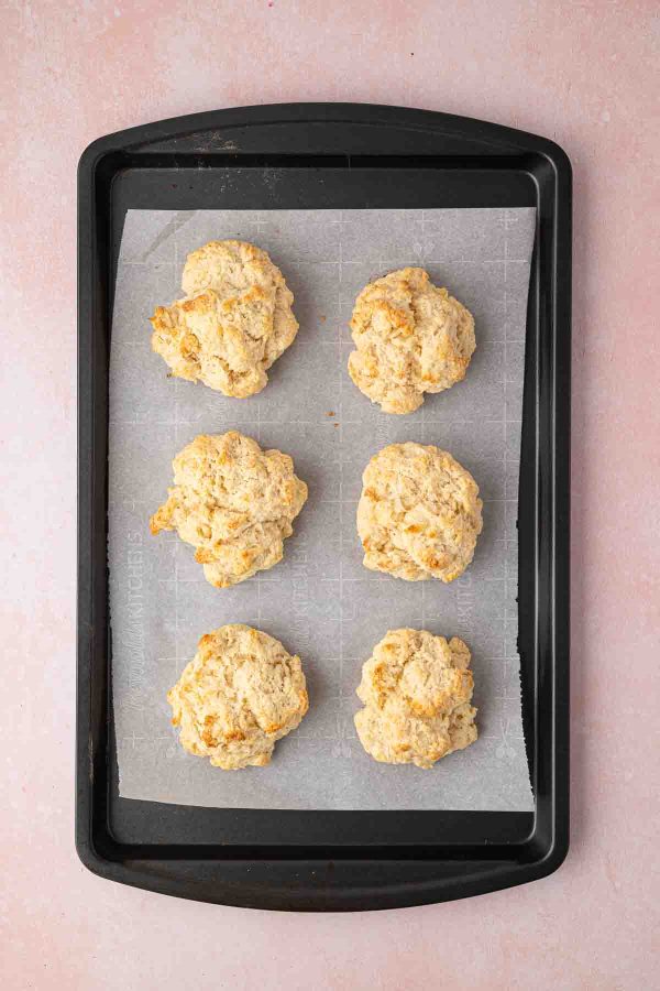 A black baking tray lined with parchment paper holds six baked drop biscuits, arranged in two columns of three, on a light pink surface.