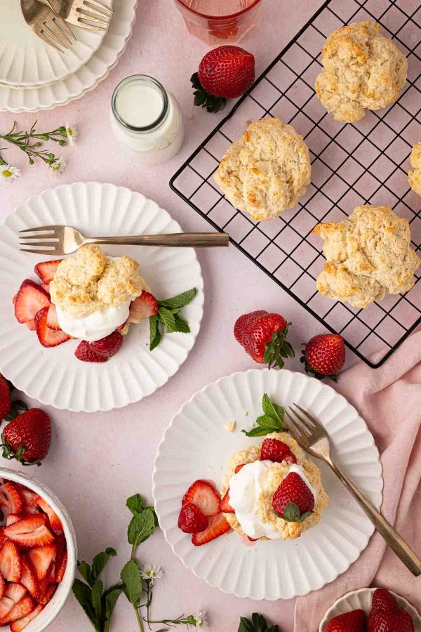 Plates of strawberry shortcake with whipped cream, fresh strawberries, and biscuits are arranged on a pink surface with a wire rack, milk, and utensils.