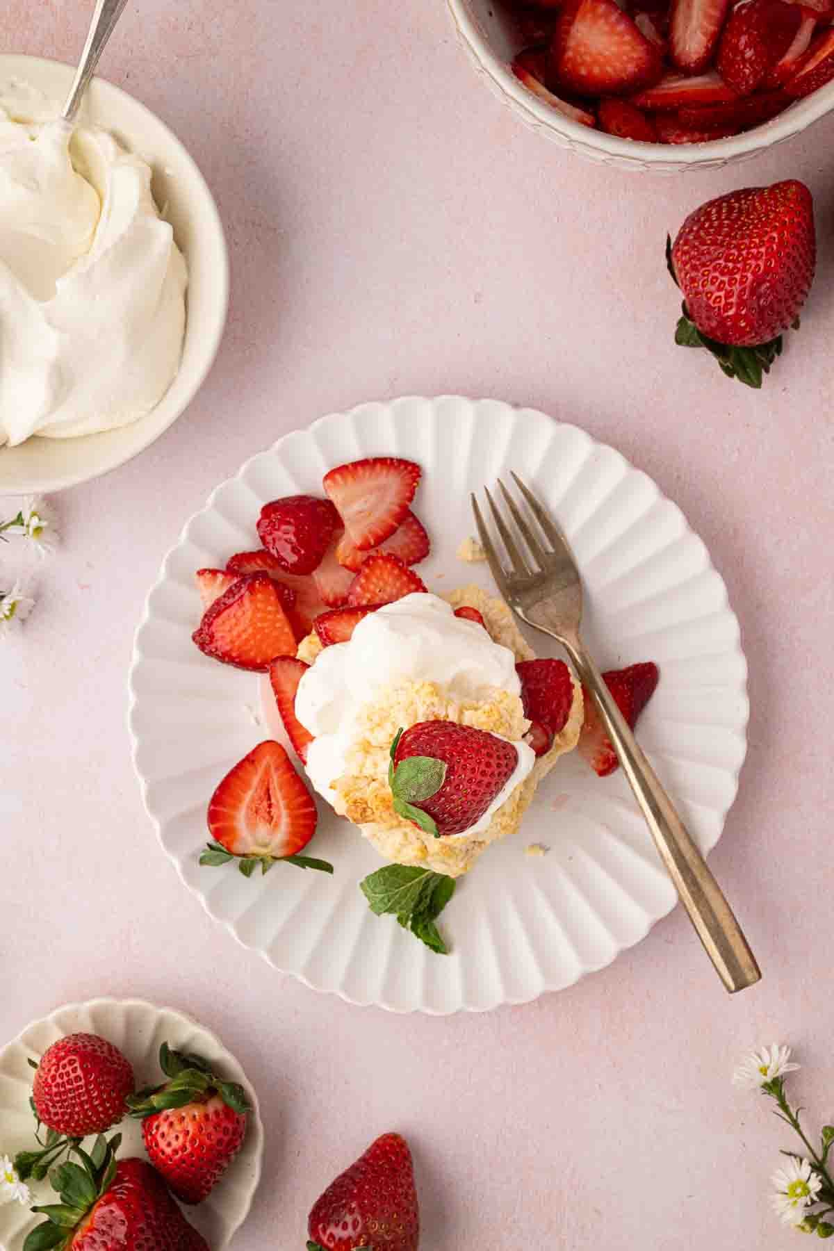 A plated dessert with whipped cream, sliced strawberries, and a mint leaf, surrounded by bowls of whipped cream, strawberries, and a fork on a light pink surface.
