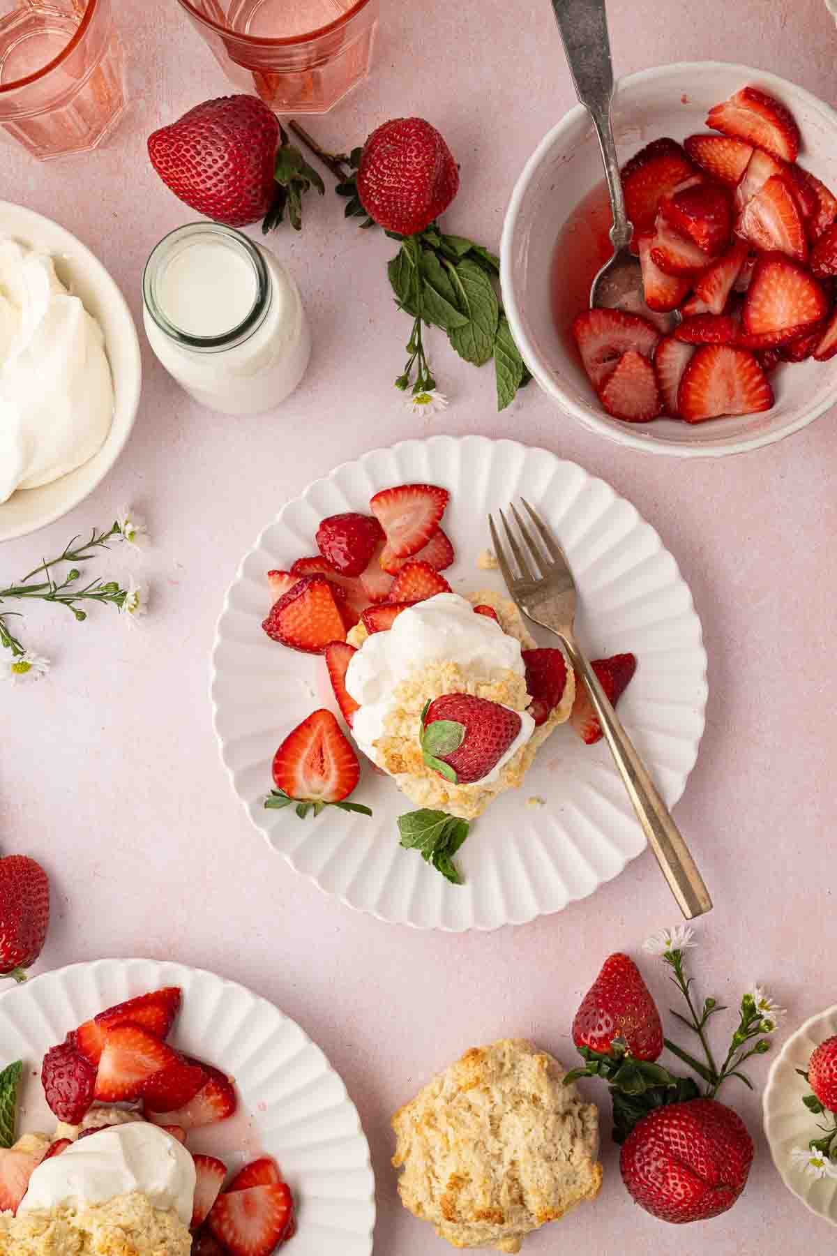 A plate with strawberry shortcake topped with whipped cream and mint, surrounded by fresh strawberries, a biscuit, a bowl of sliced strawberries, a bottle of milk, and glasses on a pink surface.