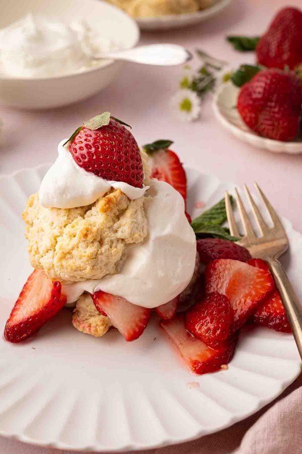 A plate with strawberry shortcake topped with whipped cream and a whole strawberry, garnished with sliced strawberries and a mint leaf, next to a fork.