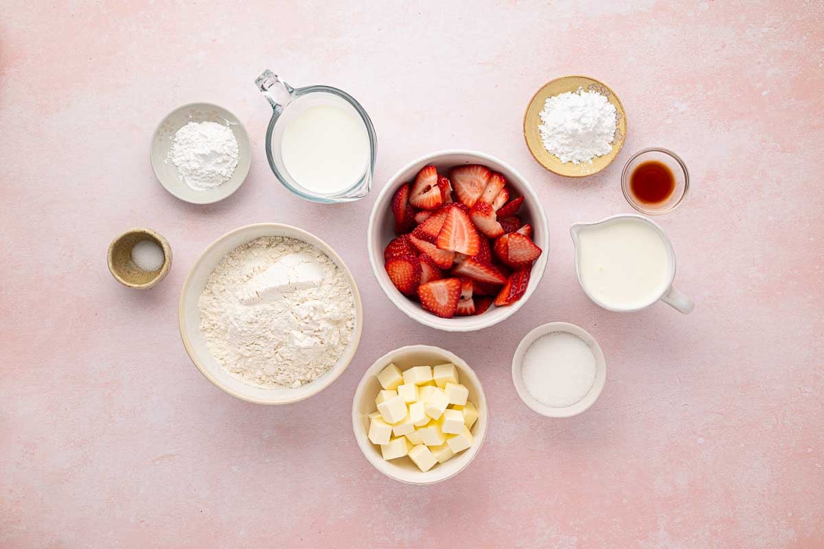 Various baking ingredients, including sliced strawberries, flour, sugar, butter, milk, cream, baking powder, cornstarch, salt, and vanilla, arranged in bowls on a pink surface.