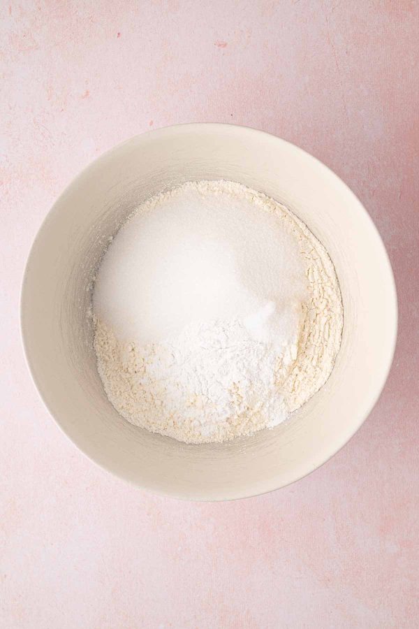 A white bowl containing a mixture of flour and granulated sugar on a light pink surface.