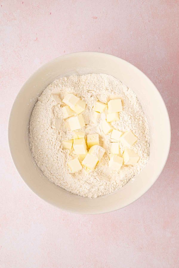 A white bowl containing flour with small cubes of butter on top, set on a light pink surface.