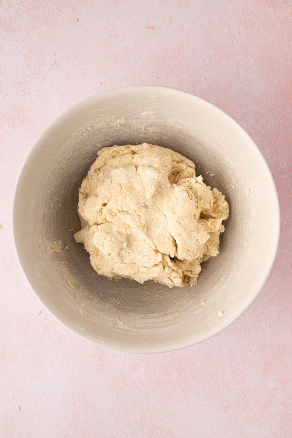 A ball of raw dough sits in a large white mixing bowl on a light pink surface.