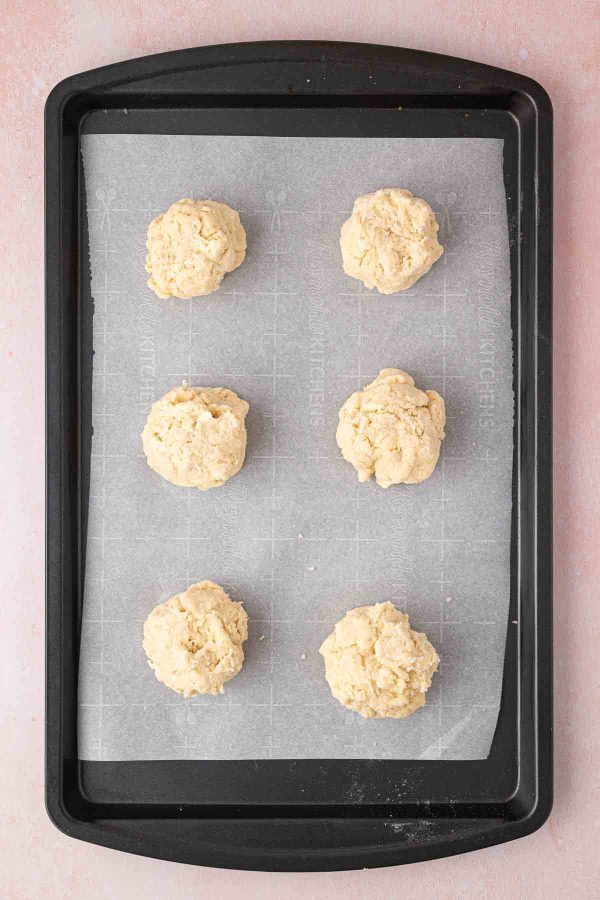 Six unbaked shortcake dough balls arranged on a parchment-lined baking sheet.
