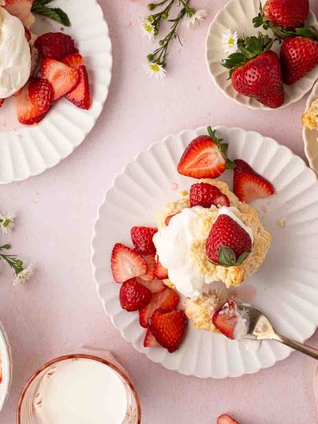 Plates with strawberry shortcakes topped with whipped cream and fresh strawberries, surrounded by whole strawberries, sliced strawberries, flowers, and a glass of milk on a pink surface.