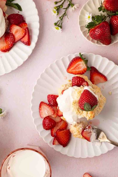 Plates with strawberry shortcakes topped with whipped cream and fresh strawberries, surrounded by whole strawberries, sliced strawberries, flowers, and a glass of milk on a pink surface.